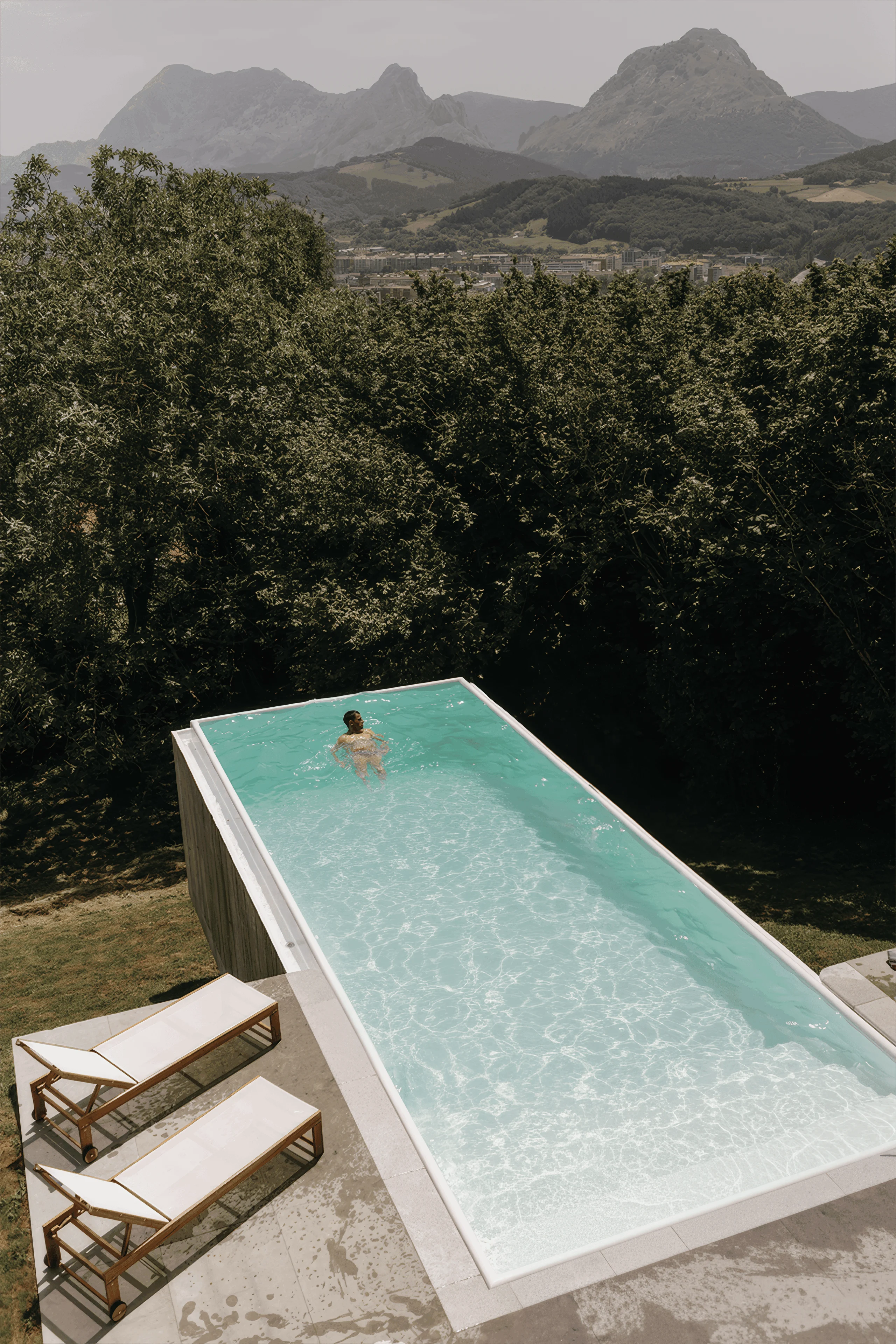 Vista elevada de la piscina con una persona en el agua, tumbonas y montañas al fondo.